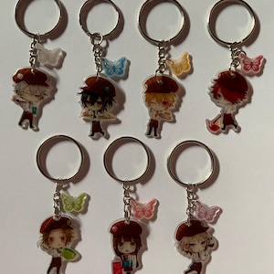 Puede incluir: Conjunto de ocho llaveros con personajes de anime con dijes de mariposa. Los personajes llevan sombreros marrones y sostienen varios objetos, como una taza de café, un libro y un pincel.