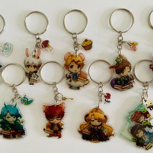Może przedstawiać: Dziesięć kolorowych breloków z postaciami z anime ze srebrnymi kółkami. Każdy brelok przedstawia inną postać z unikalnym wzorem i małym dodatkiem przyczepionym do kółka. Dodatki obejmują filiżankę do herbaty, grzyb, babeczkę, różę, klucz, ślimaka, kota i książkę.