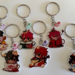 Op de afbeelding: Zeven kleurrijke sleutelhangers met cartoonfiguren in theekopjes. Elke sleutelhanger heeft een zilveren ring en een bedel met een klein ontwerp. De personages omvatten een persoon met paars haar, een persoon met rood haar, een persoon met bruin haar en een persoon met bruin haar die een hoed draagt.