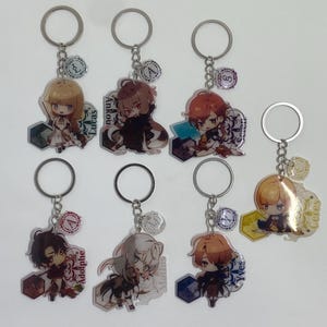 Könnte beinhalten: Sieben Schlüsselanhänger mit Charakterdesigns im Anime-Stil. Jeder Schlüsselanhänger hat einen silbernen Ring und einen kleinen Anhänger mit einem Buchstaben oder Symbol. Die Charaktere haben unterschiedliche Haarfarben und Outfits, und einige haben Namen aufgedruckt.