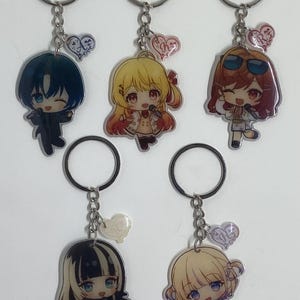 May include: Five colorful anime character keychains with silver key rings and charms. The charms are heart-shaped with the word "Love" written on them. The characters are drawn in a chibi style and are wearing different outfits.