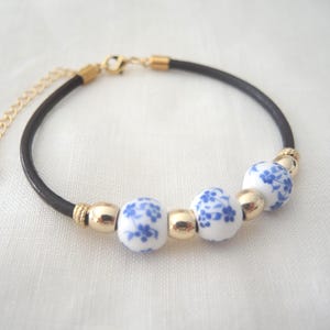 Könnte beinhalten: Ein schwarzes Lederarmband mit goldfarbenen Akzenten und weißen Keramikperlen, die mit blauen Blumenmustern verziert sind. Das Armband hat eine goldfarbene Kette mit einem herzförmigen Anhänger.