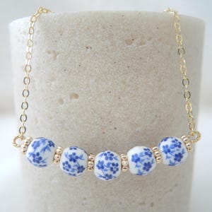 Puede incluir: Un delicado collar con cuentas de porcelana blanca con diseños florales azules. Las cuentas están ensartadas en una cadena dorada. El collar se muestra sobre un fondo neutro, resaltando su elegante diseño.