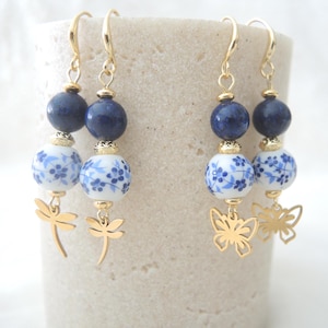 May include: Gold-coloured earrings with blue and white floral porcelain beads, dark blue beads, and gold dragonfly and butterfly charms. The earrings are suspended from gold-coloured ear wires.