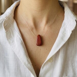 Puede incluir: Collar con colgante de piedra preciosa marrón rojizo. El colgante tiene forma alargada y lisa, suspendido de un cordón casi invisible. El collar se lleva sobre una camisa blanca abotonada.