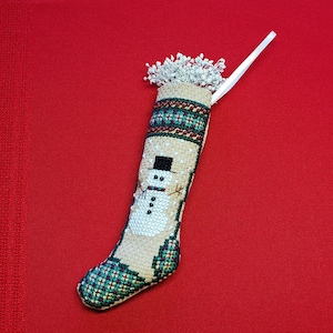 May include: A white and green Christmas stocking with a snowman design. The stocking has a white ribbon and a fluffy white pom-pom at the top.