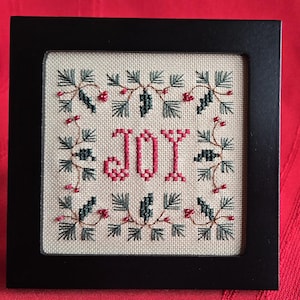 May include: A framed cross-stitch with a white background and green and red embroidery. The embroidery depicts the word "JOY" in red thread surrounded by a border of green pine needles and red berries.
