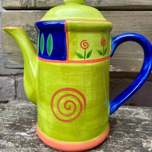 May include: A ceramic teapot featuring a bright lime green body with a blue handle, spout base, and lid knob. The design includes a large orange spiral on the front, small orange flower motifs, and green leaf shapes against a dark blue rectangular panel.