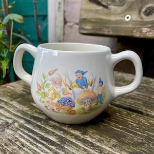 May include: A white ceramic sugar bowl with two side handles, featuring a colourful illustration of three small gnome-like figures in a forest setting, with one figure pushing a wooden wheelbarrow filled with blue berries near a large mushroom.