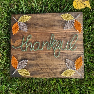 Puede incluir: Letrero de madera con la palabra "thankful" escrita con hilo verde. El letrero presenta cuatro diseños de hojas en hilo naranja, amarillo y gris. El fondo es de madera teñida de color marrón oscuro.