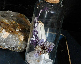 May include: A clear glass bottle with a cork stopper, containing a white feather, purple twine, dried purple flowers, and clear crystals. A large, rough-textured rock sits beside the bottle on a dark blue surface.