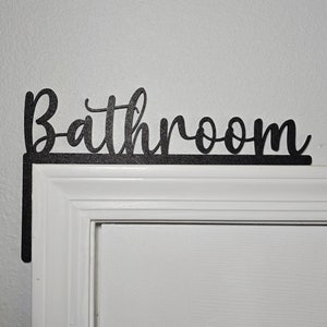 May include: Black metal sign with the word "Bathroom" in a cursive font, mounted above a white door.