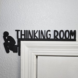May include: Black silhouette of a person sitting on a toilet with the text "Thinking Room" above it. The silhouette is attached to a white door frame.