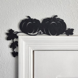 May include: Black metal pumpkin and leaf door or wall decor. The decorative piece features two pumpkins connected by a vine with leaves. The decor is mounted above a white door frame.