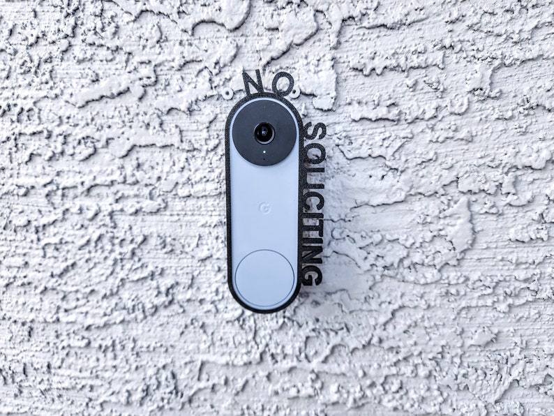 May include: A black and white sign that says "NO SOLICITING" is mounted on a white stucco wall next to a gray doorbell camera.