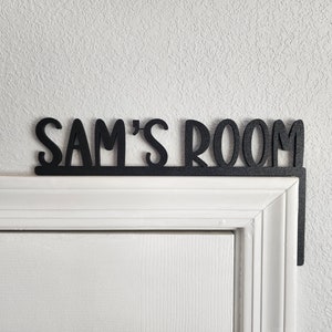 Puede incluir: Un letrero de madera negro con el texto "Sam's Room" fijado a un marco de puerta blanco.