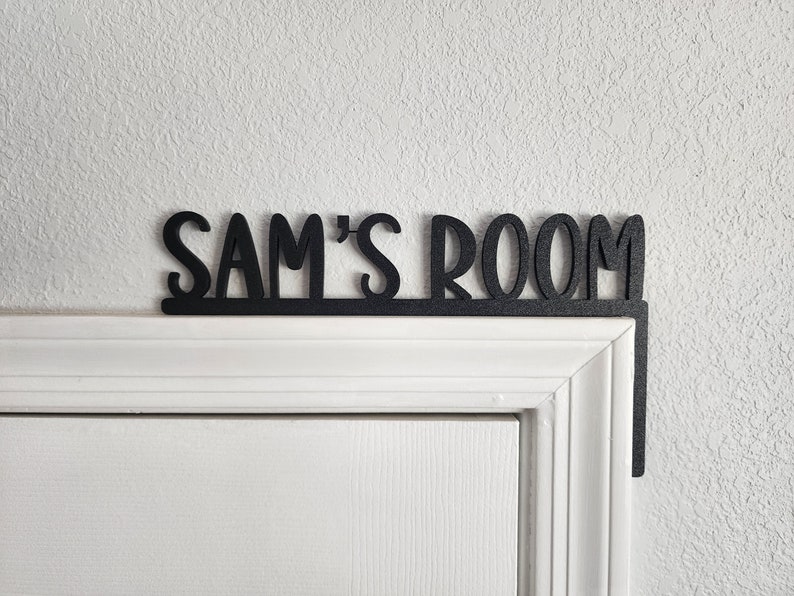 Puede incluir: Un letrero de madera negro con el texto "Sam's Room" fijado a un marco de puerta blanco.