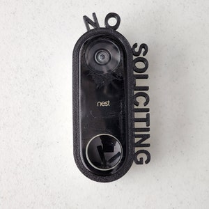 May include: A black Nest doorbell with a 3D printed sign that says "NO SOLICITING" attached to it.