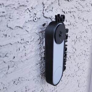 May include: A black doorbell camera with a white faceplate and a sign that says "No Soliciting".