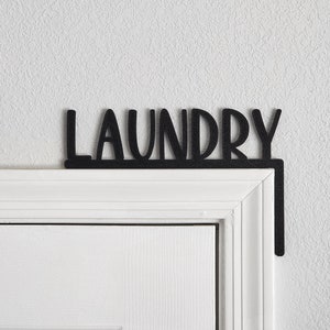 Puede incluir: Un letrero de metal negro con la palabra "LAUNDRY" en letras blancas, montado sobre un marco de puerta blanco.
