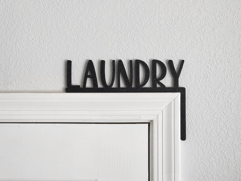 Puede incluir: Un letrero de metal negro con la palabra "LAUNDRY" en letras blancas, montado sobre un marco de puerta blanco.