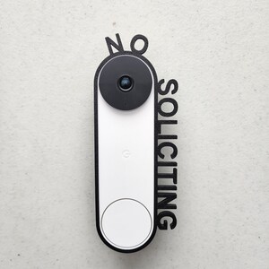 May include: A white doorbell with a black camera lens and a black frame that says "NO SOLICITING" in large letters.