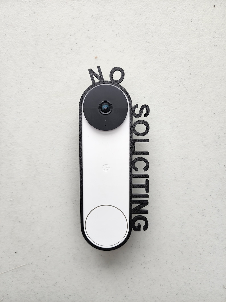 May include: A white doorbell with a black camera lens and a black frame that says "NO SOLICITING" in large letters.