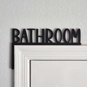 Puede incluir: Un letrero de metal negro con la palabra "BATHROOM" en may&uacute;sculas, montado sobre un marco de puerta blanco.