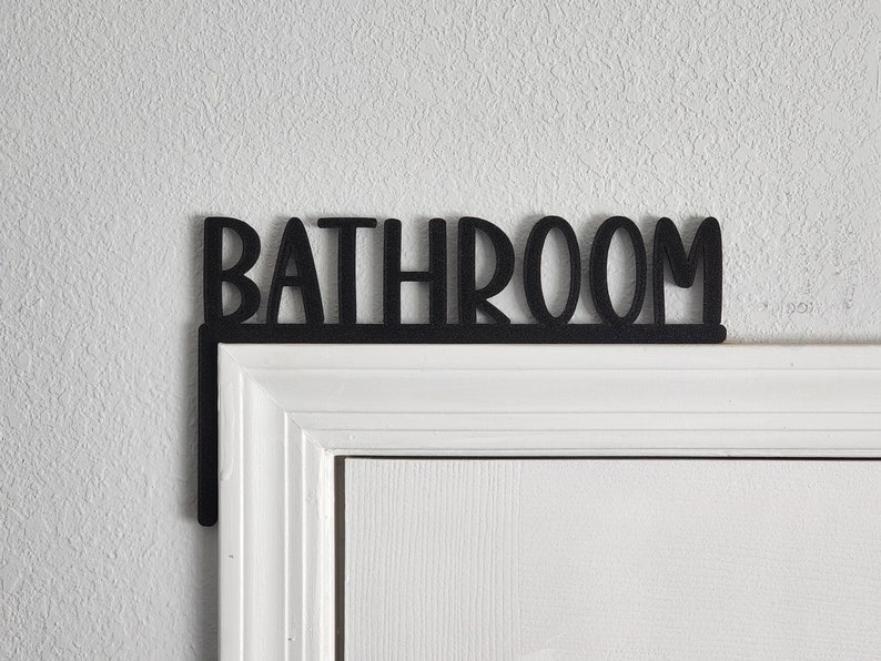 Puede incluir: Un letrero de metal negro con la palabra "BATHROOM" en may&uacute;sculas, montado sobre un marco de puerta blanco.