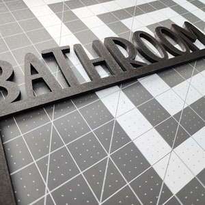 Puede incluir: Un letrero de madera negro con la palabra "BATHROOM" en may&uacute;sculas. El letrero est&aacute; inclinado y parece estar hecho de un material grueso y texturizado.