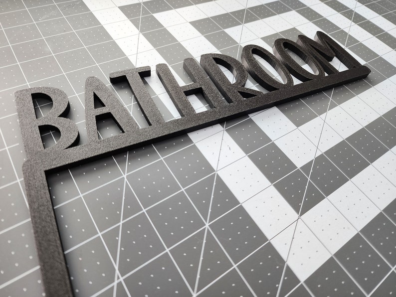 Puede incluir: Un letrero de madera negro con la palabra "BATHROOM" en may&uacute;sculas. El letrero est&aacute; inclinado y parece estar hecho de un material grueso y texturizado.