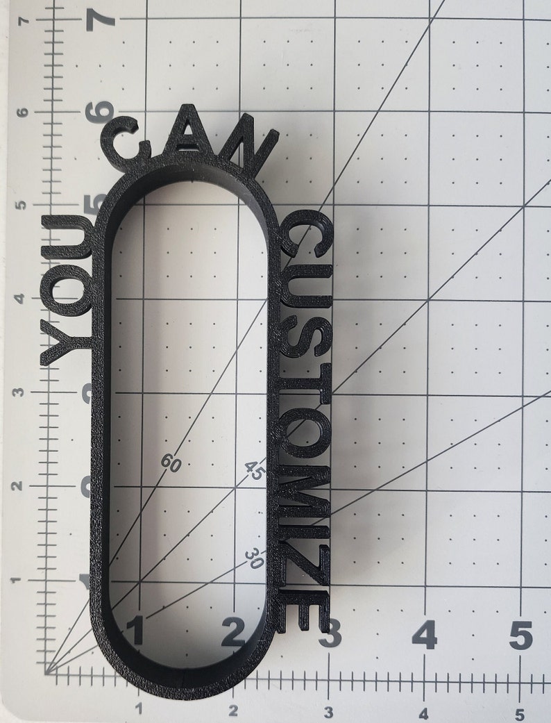 May include: A black 3D printed sign that reads "You Can Customize". The sign is shaped like a rounded rectangle with the words wrapping around the edges.