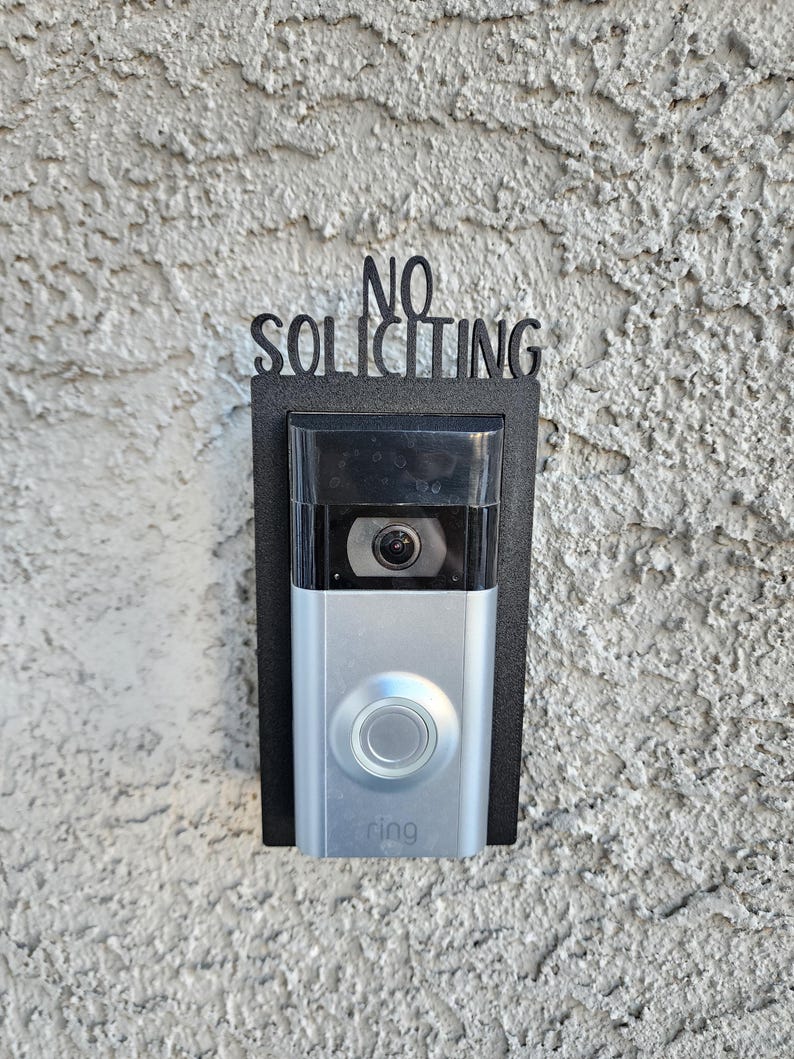 May include: A black metal sign that says "NO SOLICITING" is mounted above a silver Ring doorbell. The doorbell is mounted on a black metal plate.