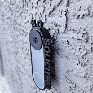 May include: A black plastic sign with the words "NO SOLICITING" printed vertically on it. The sign is mounted on a white wall next to a black doorbell camera.