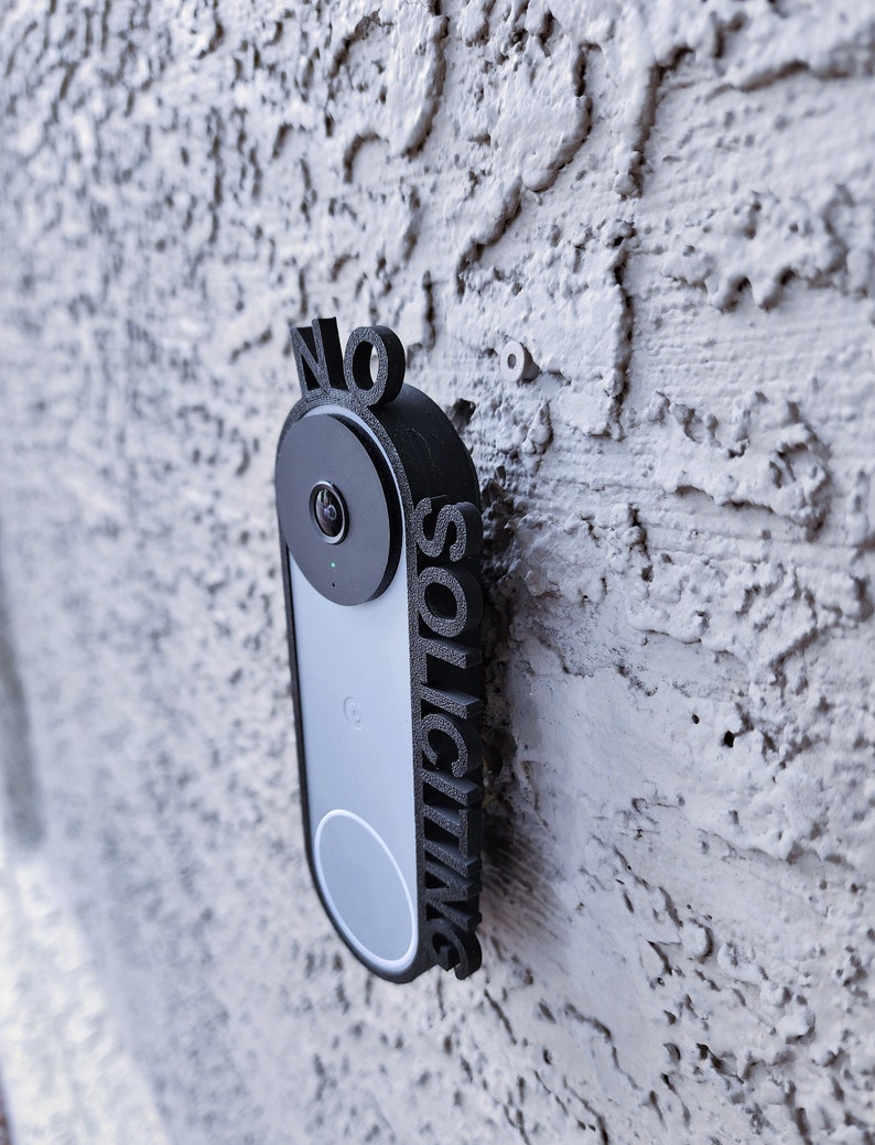 May include: A black plastic sign with the words "NO SOLICITING" printed vertically on it. The sign is mounted on a white wall next to a black doorbell camera.