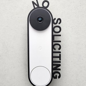 May include: A white doorbell camera with a black lens and a black frame. The frame has the words "NO SOLICITING" written vertically on it.