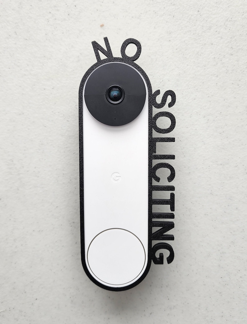 May include: A white doorbell camera with a black lens and a black frame. The frame has the words "NO SOLICITING" written vertically on it.