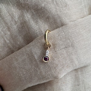 May include: A gold hoop earring featuring a dangling charm with a small rectangular silver-toned bar set with two tiny clear stones, ending in a circular bezel-set deep pinkish-red gemstone. The jewelry piece is displayed against a textured fabric.