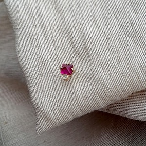 May include: A small gold stud earring featuring a square-cut vibrant pink gemstone at the top and a tiny, clear round accent stone positioned directly below it, set in a polished gold metal frame that secures the stones in place.