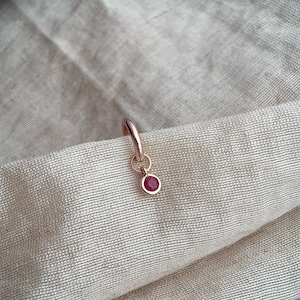May include: A rose gold hoop earring featuring a small, circular pendant with a single, round, faceted deep pink gemstone set in the centre. The metal has a polished finish, and the design is simple, consisting of a thin hoop and a charm.
