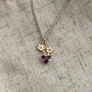 May include: A delicate necklace featuring a pendant with two small gold-colored flowers with red centers above a triangular, dark red gemstone. The chain is silver-colored, and the pendant is centered.