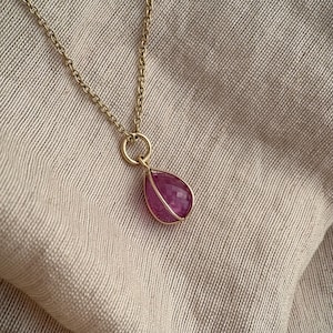 May include: A gold chain necklace featuring a teardrop-shaped, faceted pink gemstone pendant. The stone is encased in a delicate gold wire cage frame that connects to the chain via a small circular jump ring, creating a simple pendant design.