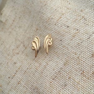 May include: A pair of gold-coloured wing-shaped earrings. Each earring has a curved, layered design with a small, circular detail at the base. The earrings are displayed on a textured, beige fabric surface.