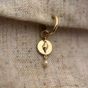 May include: A gold-colored earring with a small, round pendant featuring a seashell design. A small pearl dangles from the pendant. The earring is set against a neutral fabric background.