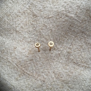 May include: A pair of small, gold-coloured stud earrings. Each earring features a circular design with a small stone in the centre. They are presented on a textured, light beige fabric. These delicate earrings would make a subtle accessory.