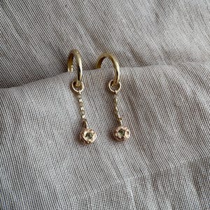 May include: A pair of gold hoop earrings with dangling chain and textured, round charms. The earrings are displayed on a neutral-toned fabric, showcasing their delicate design and warm metallic colour. The earrings are a stylish accessory.
