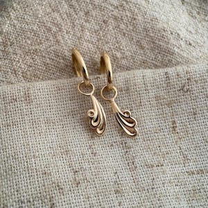 May include: A pair of gold-coloured hoop earrings with a decorative, stylised feather design. The earrings are suspended from the hoops and are displayed against a neutral, textured background. The earrings are made of a shiny, reflective metal.