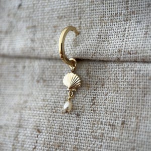 May include: A gold-coloured earring featuring a seashell charm and a small pearl. The earring is a hoop style with a hinged closure. The seashell charm has detailed ridges, and the pearl is a small, round shape.