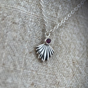 May include: A silver necklace with a delicate fan-shaped pendant featuring a small red gemstone.