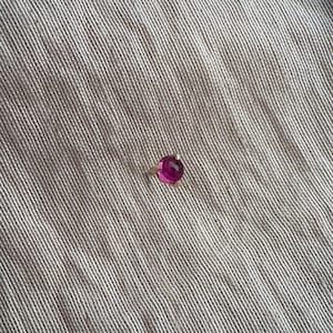 May include: A single round, faceted, vibrant magenta gemstone set in a simple gold-toned metal prong setting. The small, circular stone features a polished surface that reflects light, creating a bright, saturated pink appearance against the fabric.